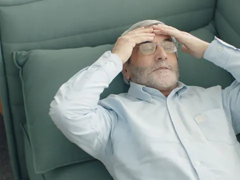 A tired older man in an armchair, hands on his head