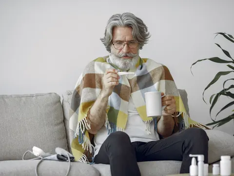 An older man with a blanket, a thermometer and a hot drink on the sofa