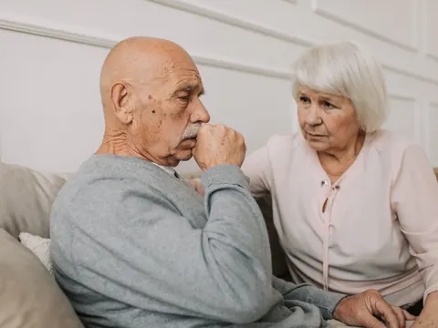 An older man coughing on the sofa, his wife beside him
